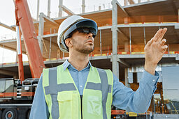 Futuristic Architectural Engineer Wearing Augmented Reality Headset and Using Gestures to Control Commercial / Industrial Building Construction Site. In the Background Skyscraper Formwork Frames and Industrial Crane Schlagwort(e): ar, architect, architecture, augmented, building, business, city, civil, construction, design, designer, development, device, engineer, engineering, future, futuristic, gesture, glasses, goggles, headset, hi-tech, house, industrial, industry, innovation, internet, investor, man, mixed reality, modern, networking, outdoors, person, project, property, real estate, reality, simulation, site, tech, technology, urban, using, virtual, virtual reality, vr, wearable, wearing, work