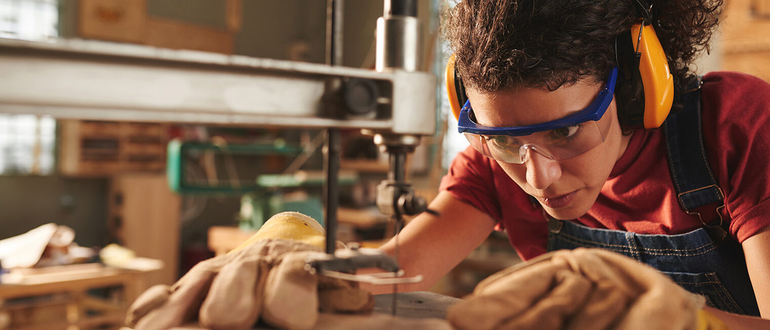 Close-up view of young concentrated female carpenter in safety glasses and ear defenders drilling hole in wooden plank with drill press Schlagwort(e): closeup, woman, female, curly, concentration, professional, carpentry, joinery, workshop, working, occupation, woodworking, processing, tool, equipment, machinery, drill, hole, press, wood, wooden, plank, board, timber, craft, craftsman, handyman, industry, manufacturing, instrument, protective, glasses, ear, defender, safety