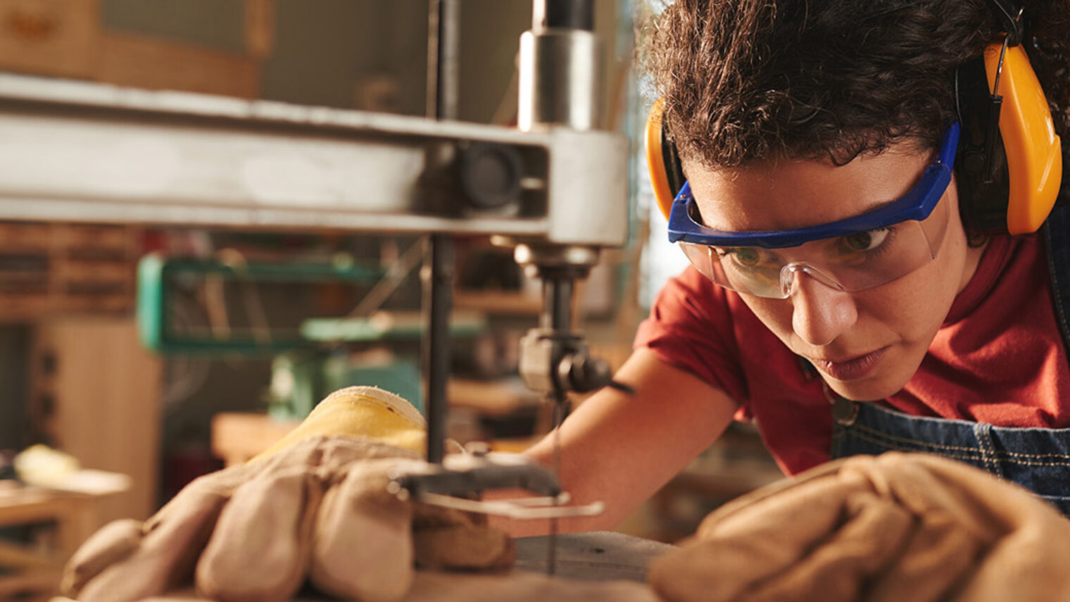 Close-up view of young concentrated female carpenter in safety glasses and ear defenders drilling hole in wooden plank with drill press Schlagwort(e): closeup, woman, female, curly, concentration, professional, carpentry, joinery, workshop, working, occupation, woodworking, processing, tool, equipment, machinery, drill, hole, press, wood, wooden, plank, board, timber, craft, craftsman, handyman, industry, manufacturing, instrument, protective, glasses, ear, defender, safety