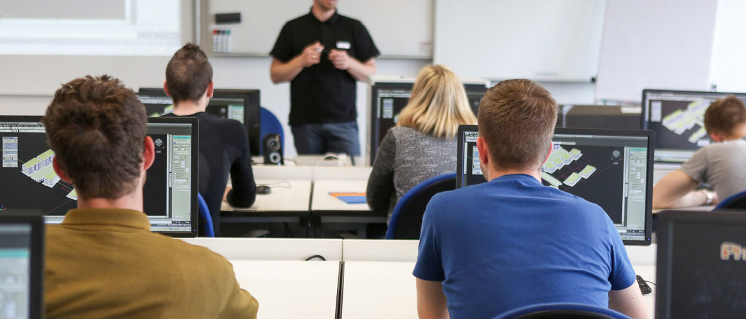 Teilnehmende eines Seminares sitzen in einem EDV Raum und schauen den Dozenten an Auszubildende nehmen an einem Seminar teil und sitzen in einem EDV Raum